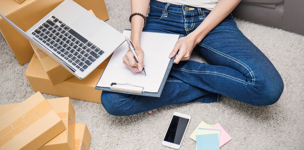 Woman taking inventory or writing notes on pad in front of laptop sitting on floor cross-legged
