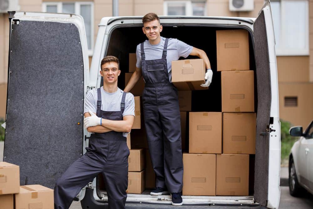 Two young workers standing in front of moving van with boxes
