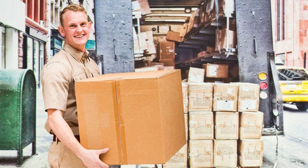 Young worker in uniform standing in front of moving truck holding a box smiling
