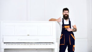 Man with beard and mustache, worker in overalls lean on piano, white background. Loader shows thumb up gesture. Courier delivers furniture in case of move out, relocation. Delivery service concept.