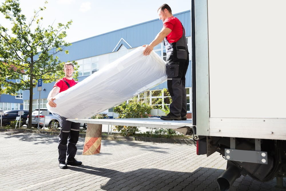 Young Male Movers Unloading The Wrapped Mattress From Moving Truck