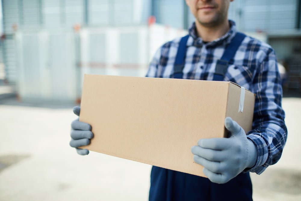 Close-up of moving company worker in gloves holding cardboard box and carrying it at container storage area