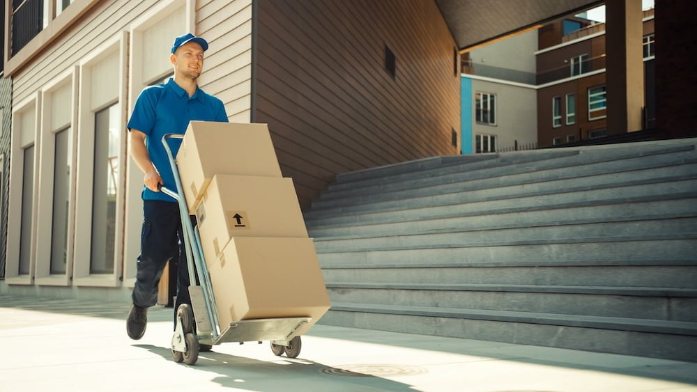 Happy Young Delivery Man Pushes Hand Truck Trolley Full of Cardboard Boxes and Packages For Delivery. Professional Courier Working Efficiently and Quickly. In the Background Stylish Modern Urban Area
