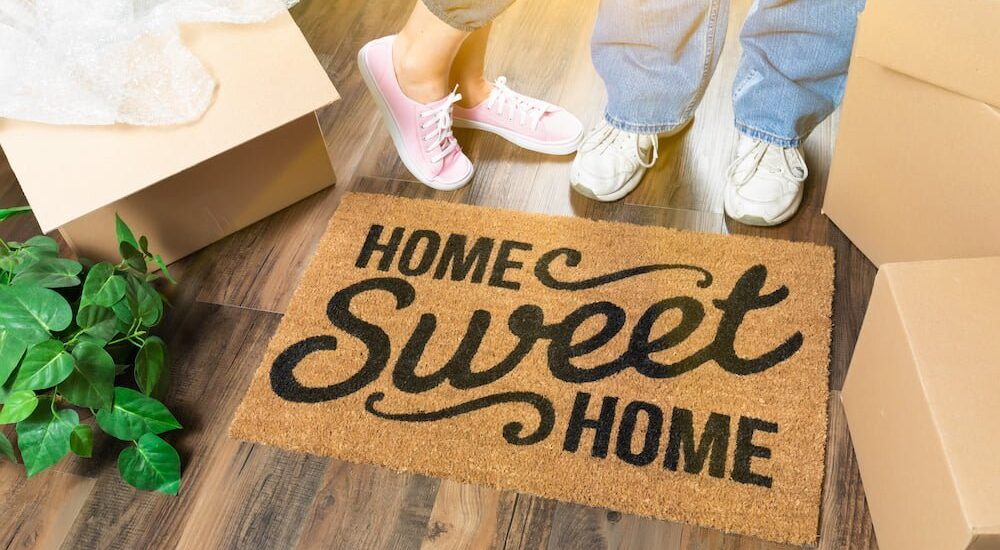 Man and Woman Standing Near Home Sweet Home Welcome Mat, Moving Boxes and Plant.