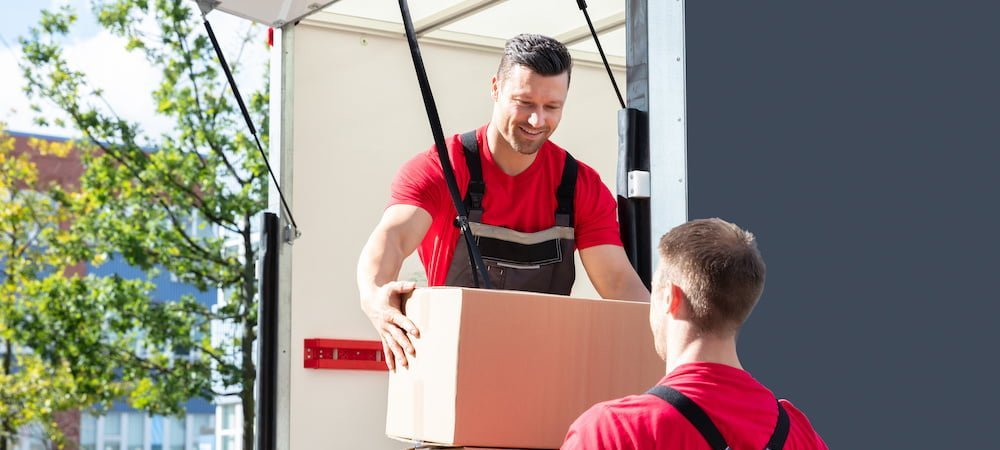 Smiling Young Male Mover Unloading In A Moving Van And Passing A Cardboard Boxes