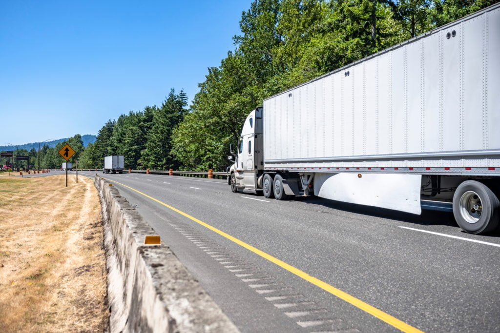 White semi-truck traveling on an open highway through a wooded area with blue skies in the background