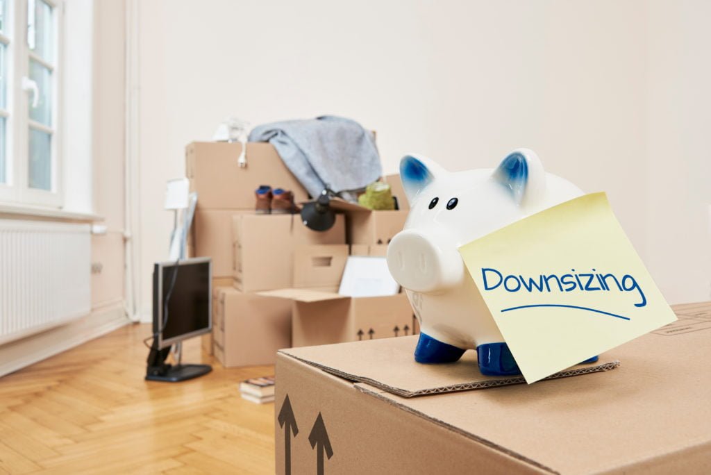 A small piggy bank sits on top of a moving box with a note attached that reads "Downsizing." Packed moving boxes sit in the background of an empty room.