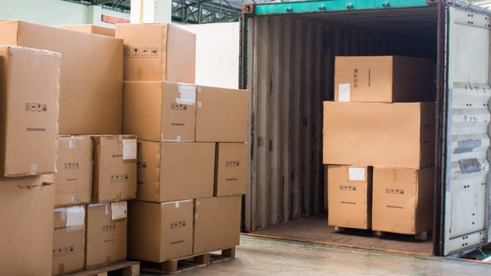 Stacked boxes in a warehouse stand next to an open moving container packed with similar boxes