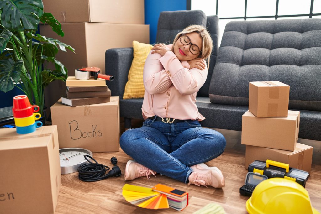 A woman hugs herself while smiling, surrounded by moving boxes in an in an indoor residential space.